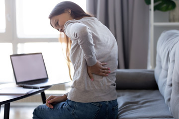 Young woman feeling pain in spine back after sedentary computer work sitting in bad posture on sofa at home, tired girl rubbing backache tensed muscles suffers from lower lumbar kidney ache, backpain