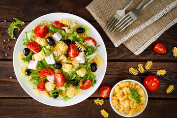 Italian pasta salad with fresh tomato, cheese, lettuce and olives on wooden background. Mediterranean cuisine. Cooking lunch.  Healthy diet food