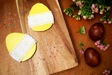 Handmade yellow easter egg out of paper on wooden background with multicolored sweet sprinkles and pink flowers and red easter eggs.