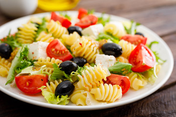 Italian pasta salad with fresh tomato, cheese, lettuce and olives on wooden background. Mediterranean cuisine. Cooking lunch.  Healthy diet food