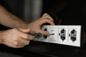 Close-up of an electrician's hand with a screwdriver disassembling a white electrical outlet on a black glass wall