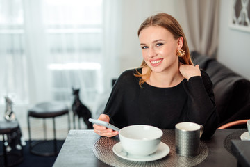 Blonde girl sit at the kitchen drink coffee and talking at her phone.