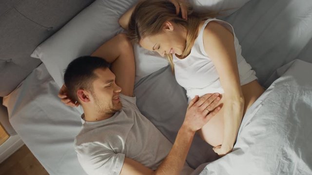 Happy Young Couple Cuddling Together in the Bed, Young Woman is Pregnant and Loving Husband Touches and Caresses Her Belly Tenderly. Top View Camera.