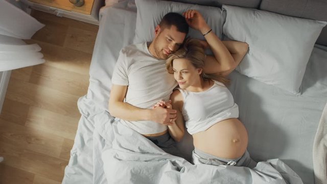 Happy Young Couple Cuddling Together in the Bed, Young Woman is Pregnant and Loving Partner Touches and Caresses Her Belly Tenderly. Sun Shines. Top Down Zoom out Camera Shot .