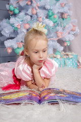 Fototapeta premium Portrait of cute european little blonde princess girl with crown in beautiful dress lying on the floor and reading book in decorated studio in christmas location with toys and christmas tree