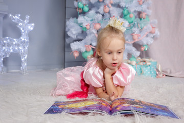 Fototapeta premium Portrait of cute european little blonde princess girl with crown in beautiful dress lying on the floor and reading book in decorated studio in christmas location with toys and christmas tree