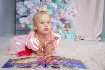 Fototapeta premium Portrait of cute european little blonde princess girl with crown in beautiful dress lying on the floor and reading book in decorated studio in christmas location with toys and christmas tree