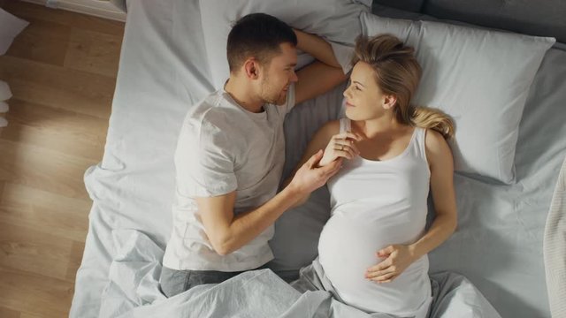 Happy Young Couple Cuddling Together in the Bed, Young Woman is Pregnant and Loving Husband Touches and Caresses Her Belly Tenderly, They Kiss. Top Down, Zoom In, Dreamy Fade Out Camera Shot.