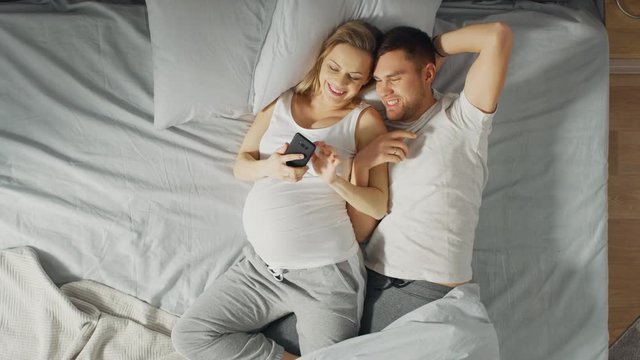 Loving Young Couple Spending Morning in Bed, Pregnant Young Woman Shows Her Partner Something on a Touchscreen Smartphone. Searching for Baby Clothes, Browsing Through Social Media, Posting Pictures.