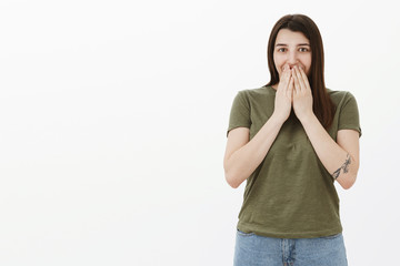 Oh my, what in there. Surprised and excited young charming girl with tattoo smiling holding hands on lips from amazement of unexpected gift grinning with eyes thrilled and delighted over gray wall