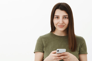 Girl taking notes in smartphone looking ready and positive at camera with tender cute smile as waiting for command grinning standing carefree and shy against gray background with mobile phone in hands
