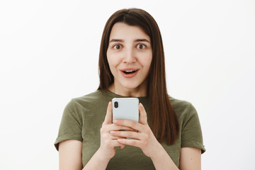 So cool best phone ever. Portrait of impressed and fascinated happy speechless brunette in 20s holding smartphone and smiling astonished at camera as excited of cellphone features over gray wall