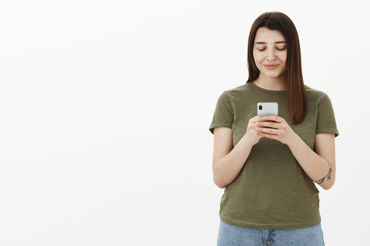 Girlfriend additing pictures on smartphone while waiting for date in cafe, standing delighted and happy over white background smiling at mobile phone screen, texting or messaging over gray wall