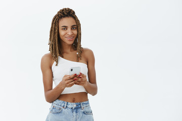 Smartphone is all you need. Portrait of stylish good-looking african american woman with cool yellow dreadlocks in white top smiling joyfully holding cellphone texting against grey wall