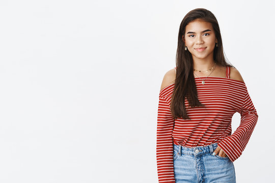 Friendly-looking stylish young indian teenage girl in striped blouse and jeans standing against gray background smiling shy at camera as holding one hand in pocket, getting know new classmates
