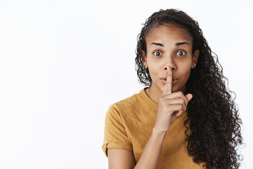 Fototapeta na wymiar Shh do not slip word. Portrait of thrilled and happy excited cute african-american woman with curly hair showing shush gesture with finger over mouth as hiding secret wanting make surprise