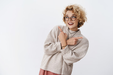 Dreamy and silly cute european blond woman pointing sideways left and right as making choice from variety of cool products smiling broadly looking up, taking decision happily over white wall