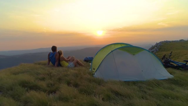 AERIAL: Flying over active couple cuddling and watching the golden sunrise above the breathtaking Alps. Carefree woman lies in her boyfriend's embrace as they sit by their tent on a beautiful evening.