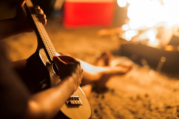 A musician woman playing ukulele guitar next to a campfire on the beach.
