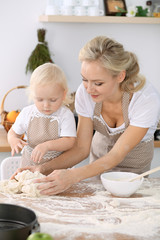 Little girl and her blonde mom in beige aprons  playing and laughing while kneading the dough in kitchen. Homemade pastry for bread, pizza or bake cookies. Family fun and cooking concept