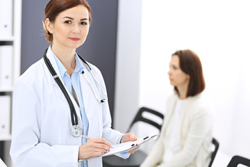 Fototapeta premium Doctor woman at work. Portrait of female physician filling up medical form while standing near reception desk at clinic or emergency hospital. Patient woman sitting at the background. Medicine concept