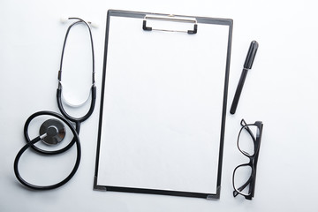 Doctor's working table with stethoscope acoustic medical device, eyeglasses and pen, blank medical record chart or test results on clipboard. Close up, copy space, background, top view, flat lay.