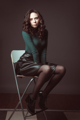 Obraz na płótnie Canvas Portrait of beautiful woman with makeup, on a chair, in green sweater and black leather skirt posing on dark chocolate background