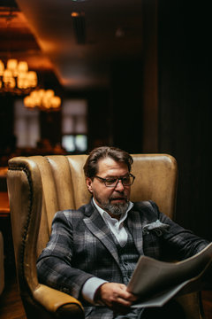 Handsome middle-aged prosperous businessman sitting in leather arm-chair in hotel lobby, wears optical spectacles is reading newspaper while waiting for his room be ready