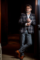 Fototapeta premium Full-size portrait of wealthy prosperous senior businessman in his fifties, dressed in stylish bespoke suit posing against dark background indoor
