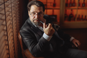 Confident pensive mature bearded male Business trainer in glasses smoking tobacco pipe, posing...