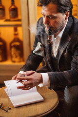 Concentrated mature start-up project developer making notes on the paper wordpad, smoking pipe with contemplating expression, sitting in the lobby of hotel