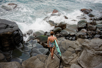 Rear view woman in the multi colored swimsuit standing with the surf on the rocks