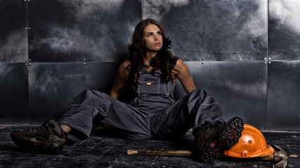 Beautiful tired sexy miner worker sitting on a floor on steel background, pickaxe and orange helmet lies nearby