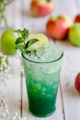 Juiced cocktail in glass and fresh fruits with leaves on white wooden background, apple and lime vitamin drink or cocktail. Closeup.