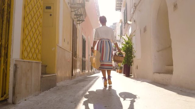 Young woman walking along alley in Ponza town island, Italy. Fashion colorful dress skirt, sunglasses, flower basket.
