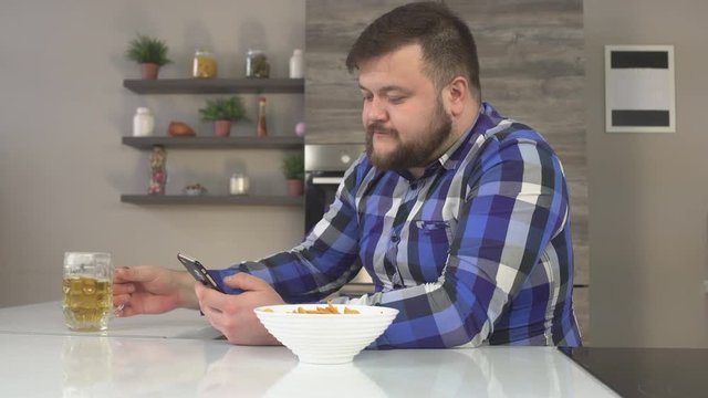 Tight caucasian man in a plaid shirt sitting in the kitchen, eating chips and drinking beer, in the hands of the phone