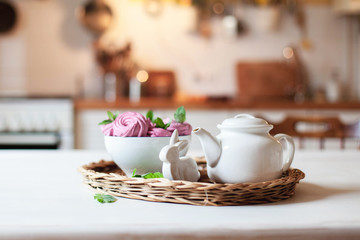 White teapot, pink cupcakes and Easter bunny are serving on straw wicker tray on table. Tea time, cozy home atmosphere hygge. Kitchen still life in warm light. Sweets with mint herbal leaves.