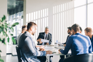 Mature grey-haired financial director reporting to his multiracial top management co-workers of income increase for this year, showing graphs with results of successful teamwork