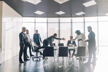 Ironic shot of caucasian businessman climbs over the office desk on his knees to reach out for a hand shake with his Arabian opponent. Reconciliation, concord and agreement concept.