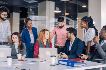 Creative multiracial business team discussing project, standing around workplace, while two co-workers with portable computers looking for needed information, sharing results to the workers