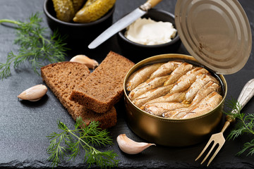Canned sprats in tin can with bread for making sandwich on dark  background.