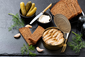 Canned sprats in tin can with bread for making sandwich on dark  background.