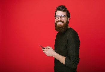 Happy bearded man in glasses typing sms adn looking at camera on red background
