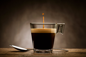 Black coffee in glass cup with teaspoon and jumping drop, on wooden table