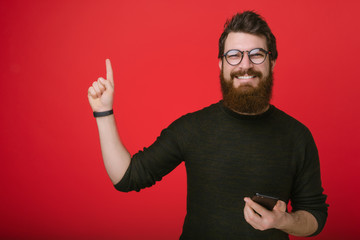 Cheerful bearded man in glasses, holding a smartphone and pointing finger at copy space