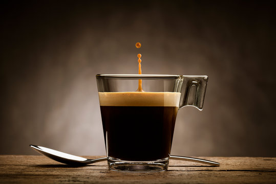 Fototapeta Czarna kawa w szklanym kubku z łyżeczką i skoki kropli, na drewnianym stole
