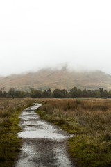 Hiking path towards a mountain covered in fog during a rainy autumn day with rain puddles near Kilchurn Castle (Scotland, United Kingdom, Europe)