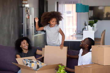 Happy child girl jump out of box play with parents in living room, excited joyful african family...
