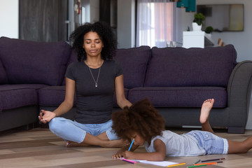 Calm african american mom doing yoga while quiet little mixed race kid daughter drawing on warm...