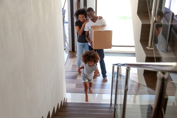 Cute african american child girl running up stairs exploring new big house moving in, happy black parents holding boxes entering own home with kid daughter enjoy relocation, family mortgage concept
